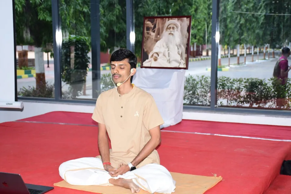 Shrungar seated in meditation on a mat, eyes closed, wearing a headset microphone